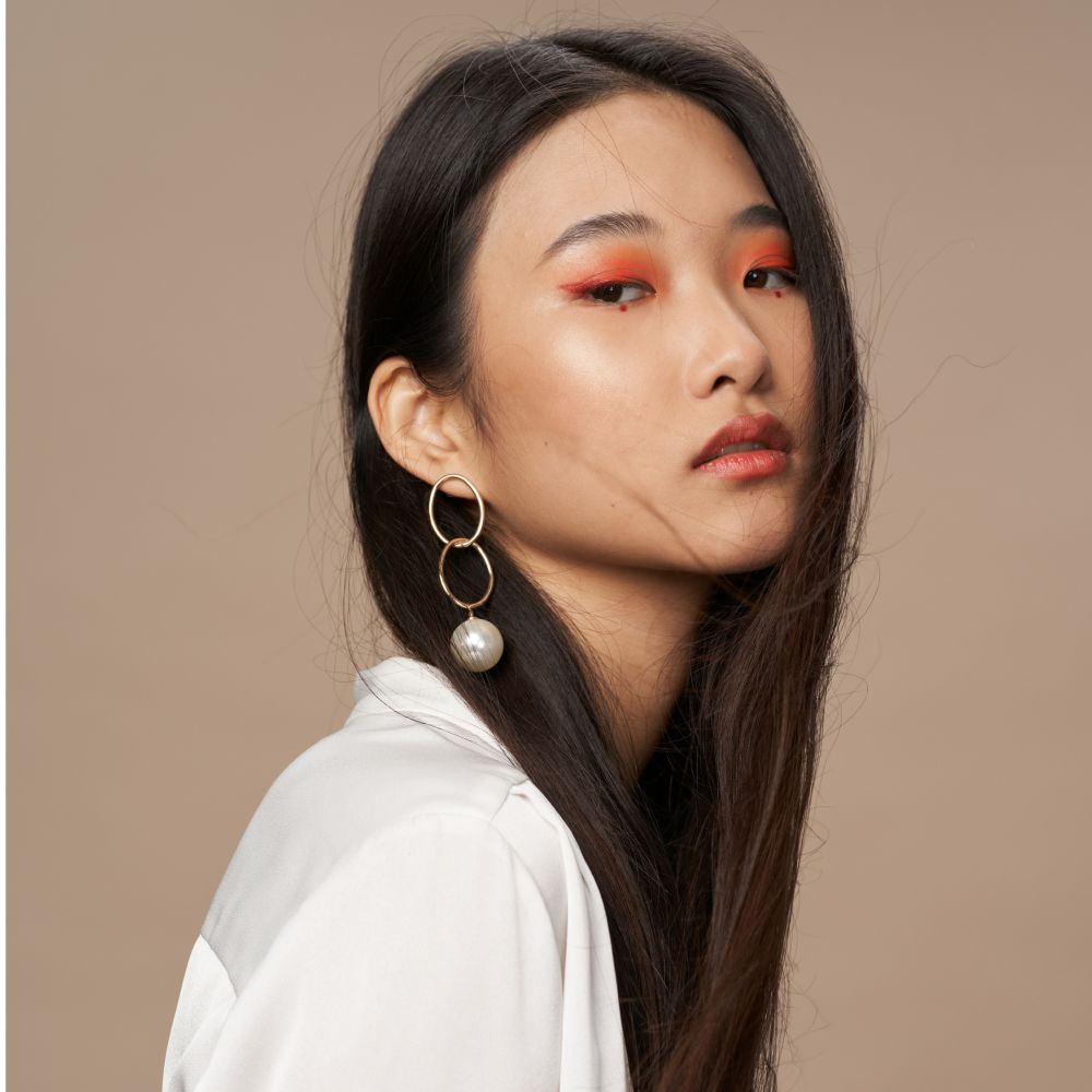 Woman with long dark hair wearing gold earrings and orange eyeshadow, against a neutral background.