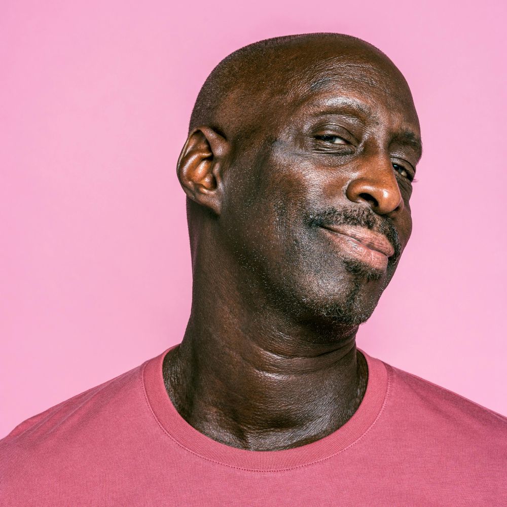 Man with a bald head and slight smile, wearing a pink shirt, against a pink background.
