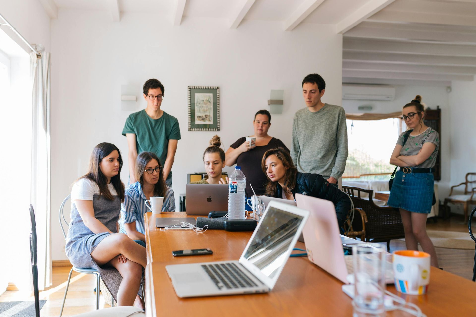Group of people around a table, looking at laptops. Indoors, casual setting. Some pointing, observing.
