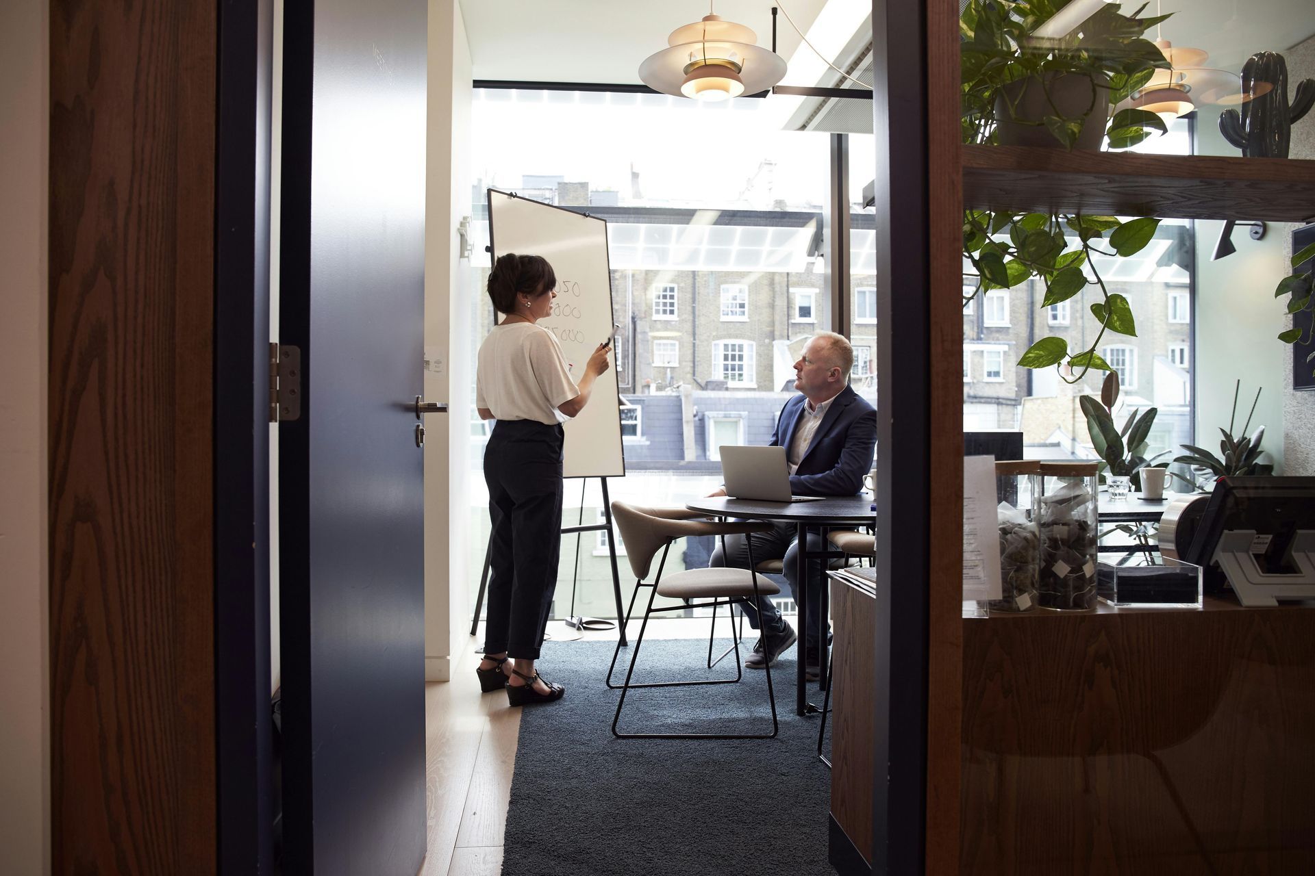 Woman presenting to a man in an office meeting room, pointing at a whiteboard.