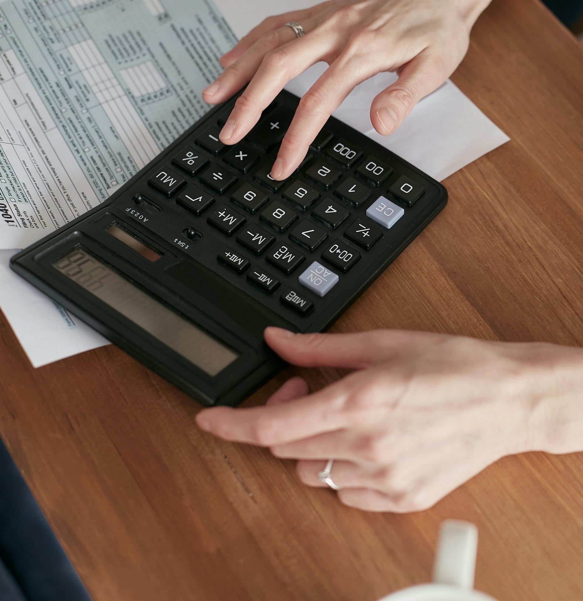 Hands using a calculator on a wooden table with paperwork; coffee cup in frame.
