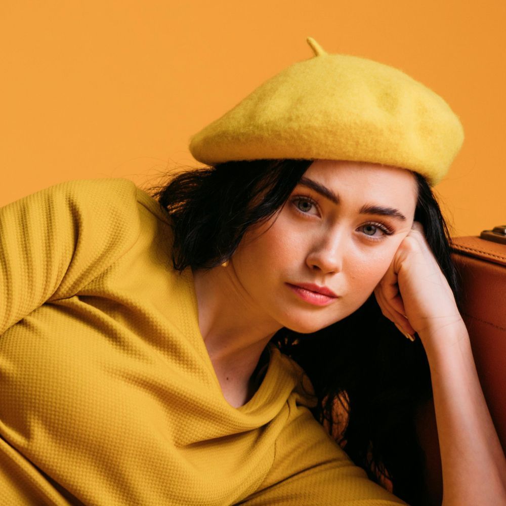 Woman in yellow beret and shirt, leaning on a brown suitcase against a yellow backdrop.
